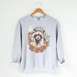 May include: A gray sweatshirt with a graphic of a raccoon sitting in a crescent moon surrounded by flowers and mushrooms.