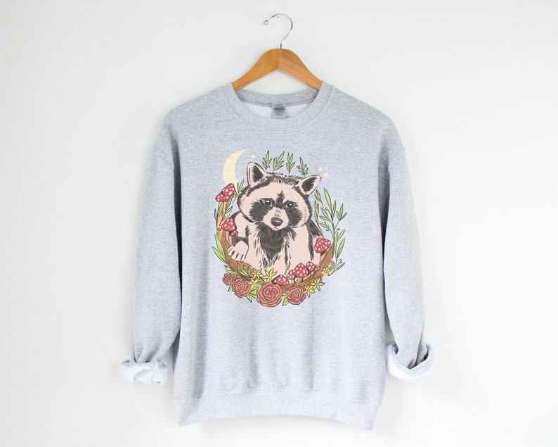 May include: A gray sweatshirt with a graphic of a raccoon sitting in a crescent moon surrounded by flowers and mushrooms.