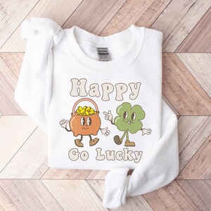 May include: White sweatshirt with a graphic of a pot of gold and a four-leaf clover with the text "Happy Go Lucky" printed on it.
