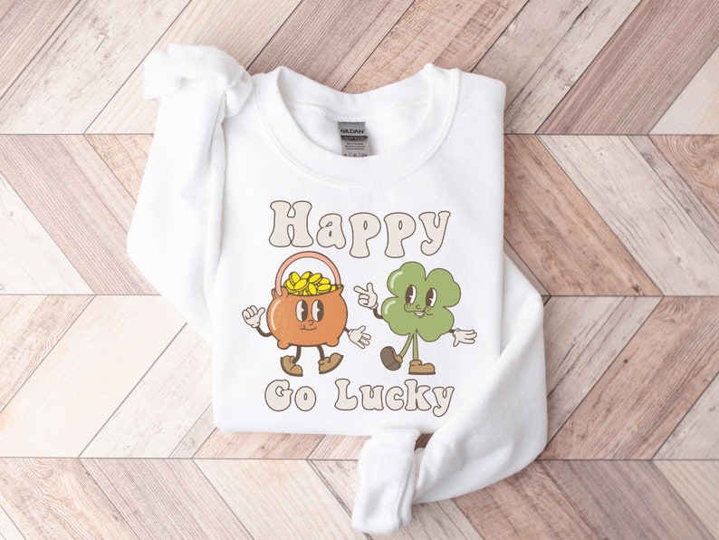 May include: White sweatshirt with a graphic of a pot of gold and a four-leaf clover with the text "Happy Go Lucky" printed on it.