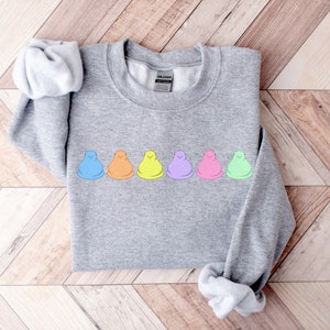 Peut inclure: Un sweat-shirt gris avec une rang&eacute;e de six guimauves en forme de poussins pastel imprim&eacute;es sur le devant. Les guimauves sont dispos&eacute;es en ligne de gauche &agrave; droite dans les couleurs suivantes&nbsp;: bleu, orange, jaune, violet, rose et vert.