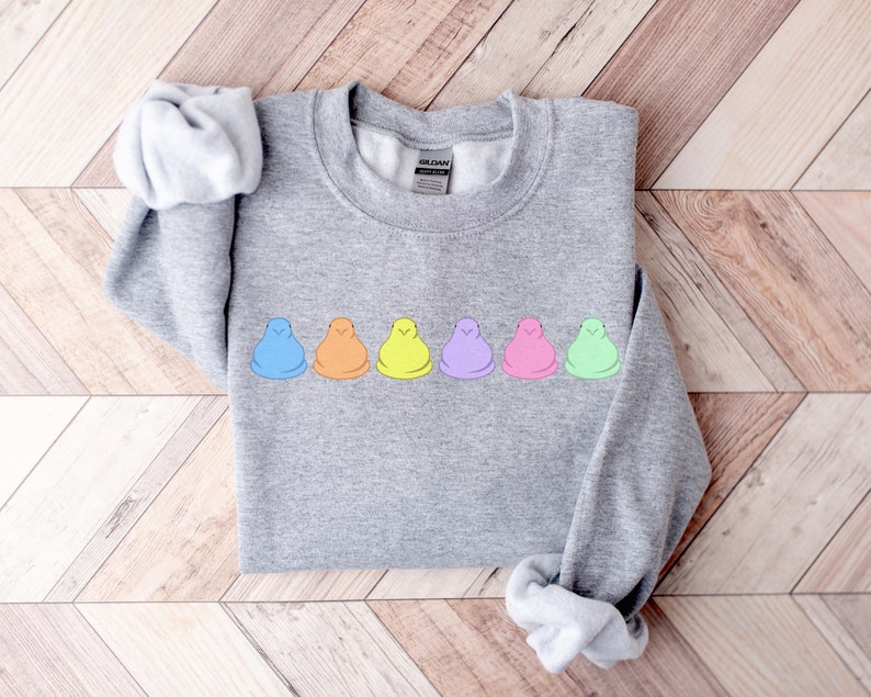 Peut inclure: Un sweat-shirt gris avec une rang&eacute;e de six guimauves en forme de poussins pastel imprim&eacute;es sur le devant. Les guimauves sont dispos&eacute;es en ligne de gauche &agrave; droite dans les couleurs suivantes&nbsp;: bleu, orange, jaune, violet, rose et vert.