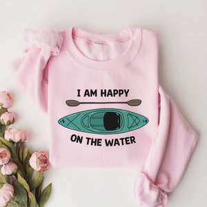 May include: Pink sweatshirt with a graphic of a kayak and paddles. The text "I am happy on the water" is printed on the sweatshirt.