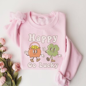 May include: Pink sweatshirt with a graphic of a pot of gold and a four-leaf clover with the text "Happy Go Lucky".
