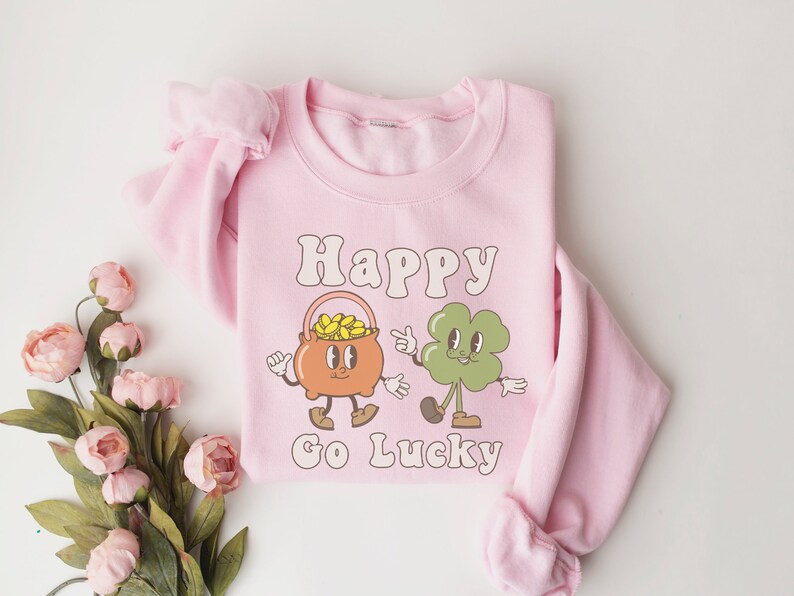 May include: Pink sweatshirt with a graphic of a pot of gold and a four-leaf clover with the text "Happy Go Lucky".