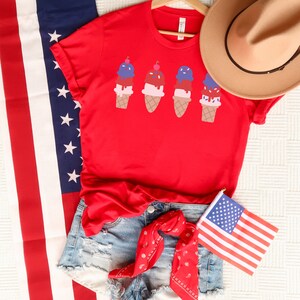 May include: Red t-shirt featuring a graphic of four ice cream cones in red, white and blue colours. The shirt is styled with denim shorts and a red bandana.