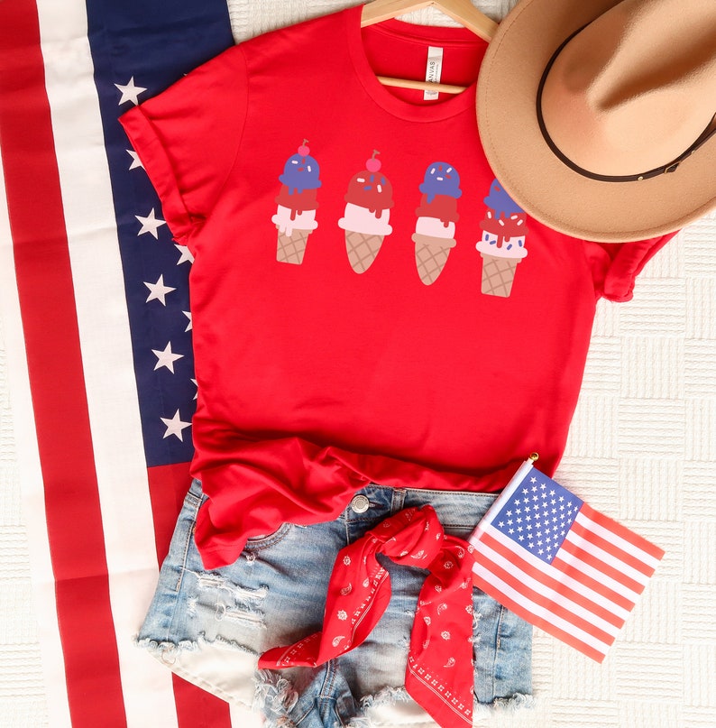 May include: Red t-shirt featuring a graphic of four ice cream cones in red, white and blue colours. The shirt is styled with denim shorts and a red bandana.