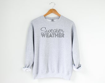 May include: A light gray sweatshirt with the words "Sweater Weather" printed in black script. The sweatshirt is hanging on a wooden hanger against a white background. The sleeves are cuffed.