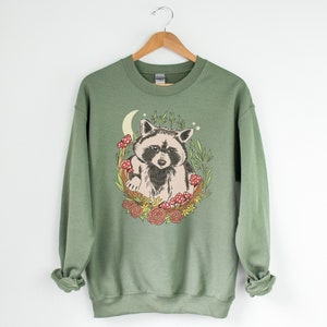 May include: A green sweatshirt with a graphic of a raccoon sitting in a crescent moon with mushrooms and flowers.