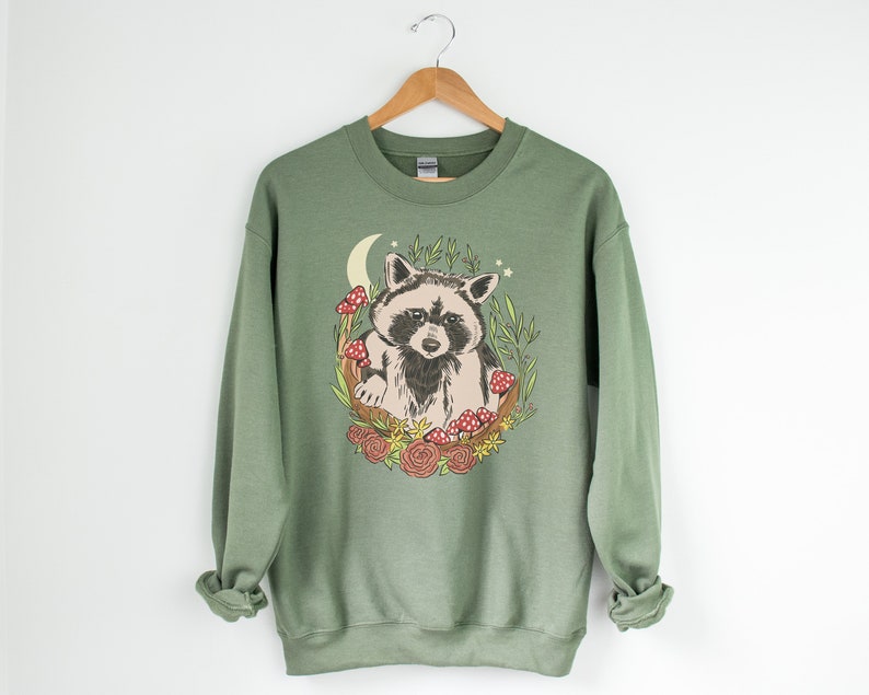 May include: A green sweatshirt with a graphic of a raccoon sitting in a crescent moon with mushrooms and flowers.