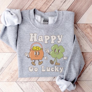 May include: A gray sweatshirt with the text "Happy Go Lucky" in a retro font. The design features a cartoon pot of gold and a four-leaf clover.