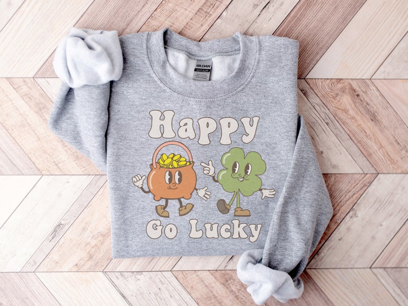 May include: A gray sweatshirt with the text "Happy Go Lucky" in a retro font. The design features a cartoon pot of gold and a four-leaf clover.