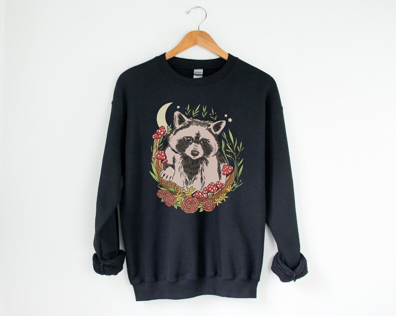 May include: Black sweatshirt with a graphic of a raccoon surrounded by mushrooms, flowers, and a crescent moon.