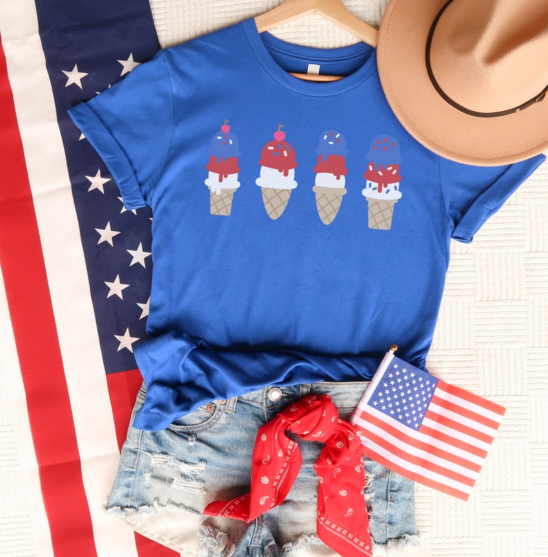 May include: A blue t-shirt featuring a design of four ice cream cones with red, white and blue scoops. The ice cream cones are topped with cherries and sprinkles. The shirt is worn with denim shorts and a red bandana.