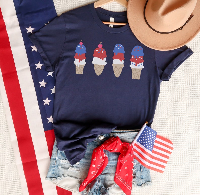 May include: A navy blue t-shirt featuring a graphic of four ice cream cones in red, white, and blue. The ice cream cones are topped with a cherry and sprinkles. The shirt is styled with denim shorts, a red bandana, and a small American flag.