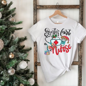 May include: White t-shirt with a Christmas-themed design that says "Santa's Favorite Nurse" with a red cross, stethoscope, bandages, and a coffee cup with a Santa hat.