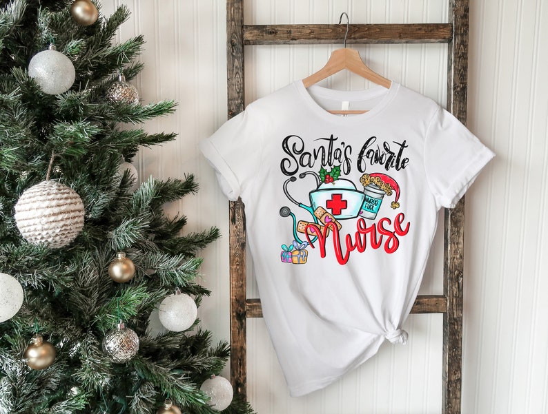 May include: White t-shirt with a Christmas-themed design that says "Santa's Favorite Nurse" with a red cross, stethoscope, bandages, and a coffee cup with a Santa hat.