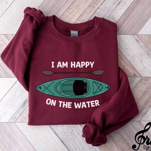 May include: Burgundy sweatshirt with the text "I am happy on the water" and a green kayak with a paddle.