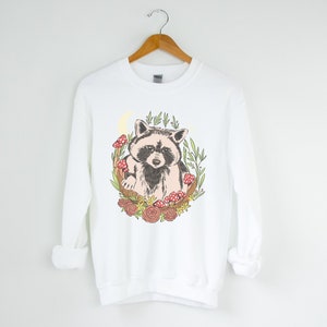 May include: White sweatshirt with a graphic of a raccoon surrounded by flowers, mushrooms, and leaves.
