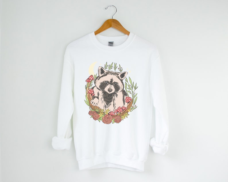 May include: White sweatshirt with a graphic of a raccoon surrounded by flowers, mushrooms, and leaves.