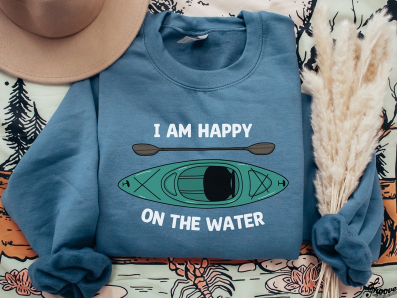 May include: A blue sweatshirt with the text "I am happy on the water" and a cartoon image of a green kayak with a paddle.