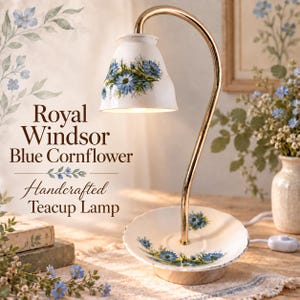 May include: A lamp featuring a white ceramic teacup shade and a matching saucer base, both decorated with blue cornflower patterns. A curved gold-tone metal arm connects the base to the shade. The text Royal Windsor Blue Cornflower Handcrafted Teacup Lamp is visible.