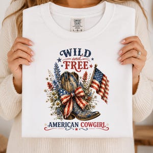 May include: White t-shirt with a patriotic design featuring a cowboy boot, American flag, and floral elements. The text reads "WILD and FREE" and "AMERICAN COWGIRL". The shirt is a Comfort Colors brand.
