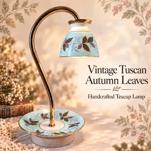 May include: A lamp featuring a curved gold-tone metal arm and a light blue ceramic teacup shade and saucer base, both decorated with a pattern of brown and purple autumn leaves. The text reads: Vintage Tuscan Autumn Leaves Handcrafted Teacup Lamp.
