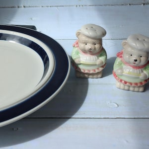 May include: A white ceramic plate with a navy blue border is next to two bear-shaped salt and pepper shakers. The shakers are dressed as chefs, wearing white hats, green jackets, and red aprons.