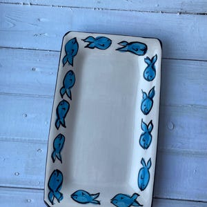 May include: Rectangular white ceramic serving platter with a black border. The platter is decorated with hand-painted blue fish motifs around the edges. A decorative piece for serving appetizers or snacks.