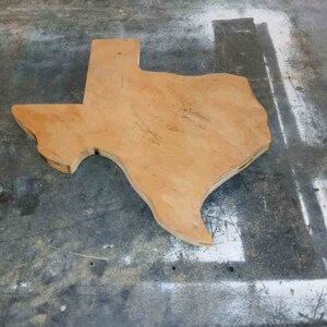 May include: A wooden cutout of the state of Texas, with a light brown color and a rough, unfinished texture. The cutout is lying on a gray, textured surface.