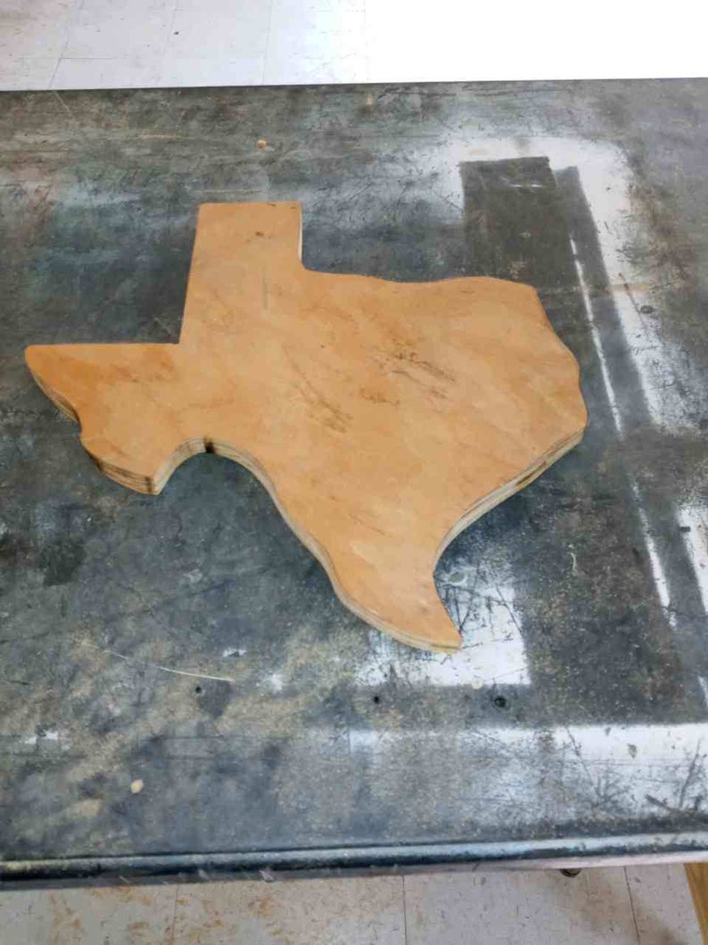 May include: A wooden cutout of the state of Texas, with a light brown color and a rough, unfinished texture. The cutout is lying on a gray, textured surface.