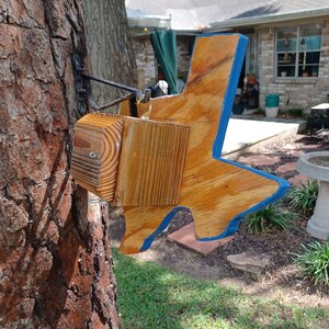 May include: A wooden bird feeder shaped like the state of Texas, painted blue around the edges, is hanging from a tree branch. The feeder is made of natural wood and has a small, square opening on the side.