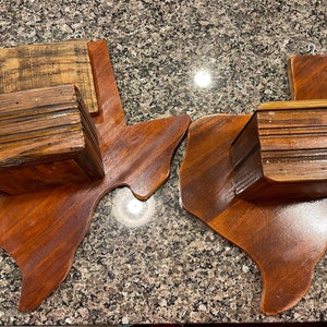 May include: Two wooden Texas state-shaped wall shelves with a rustic wood finish. Each shelf has a small wooden box attached to the top.