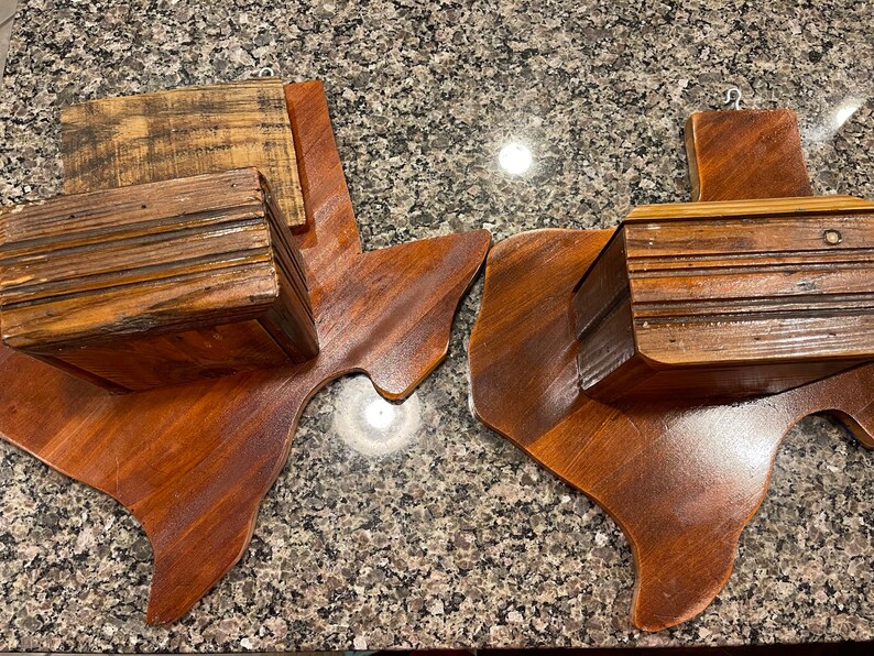 May include: Two wooden Texas state-shaped wall shelves with a rustic wood finish. Each shelf has a small wooden box attached to the top.
