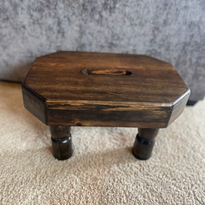 May include: A dark brown wooden stool with a square top and four legs. The stool has a slightly rounded top and a small hole in the center.