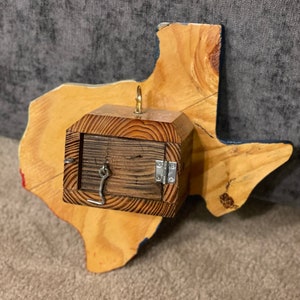 May include: A wooden box with a hinged lid and a metal latch is attached to a wooden cutout of the state of Texas. The box is painted brown and the Texas cutout is painted in the colors of the Texas flag.