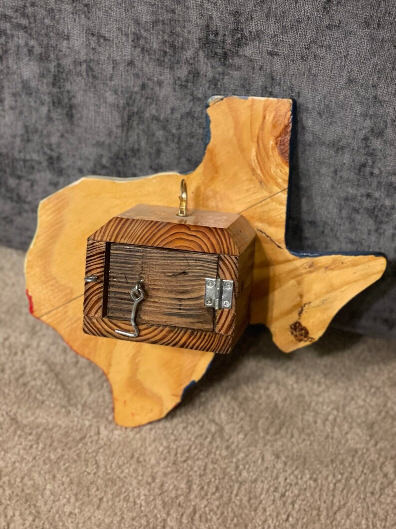 May include: A wooden box with a hinged lid and a metal latch is attached to a wooden cutout of the state of Texas. The box is painted brown and the Texas cutout is painted in the colors of the Texas flag.