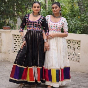 May include: Two women wearing long, embroidered dresses. One dress is black with colorful accents at the hem, the other is white. Both dresses have three-quarter sleeves and intricate embroidery around the neckline and bodice. They are standing in front of a white wall.