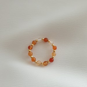 May include: A beaded ring featuring a circular design. The ring is composed of small, round beads in shades of orange, white, and gold. The beads are strung together, creating a continuous loop. The ring is set against a white background.
