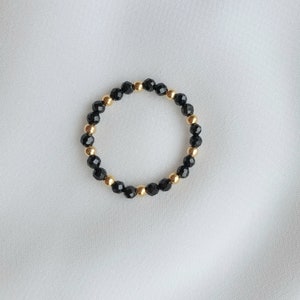 May include: A beaded ring featuring alternating black faceted beads and gold-colored round beads. The ring is set against a white background, showcasing the jewelry's design and color contrast. The ring is a stretch style.