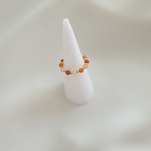 May include: A beaded ring featuring a stretch design. The ring is adorned with a mix of orange, yellow, and white beads, interspersed with small gold-colored beads. The ring is displayed on a white, cone-shaped stand against a white background.
