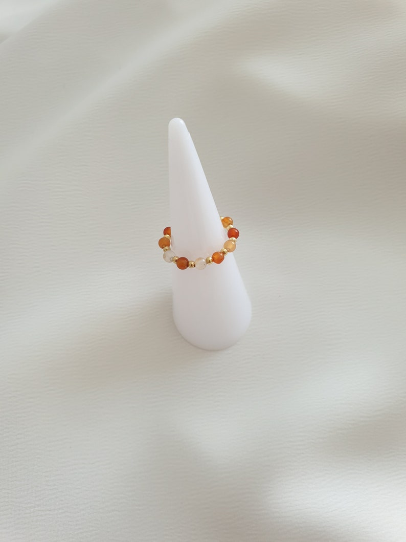 May include: A beaded ring featuring a stretch design. The ring is adorned with a mix of orange, yellow, and white beads, interspersed with small gold-colored beads. The ring is displayed on a white, cone-shaped stand against a white background.