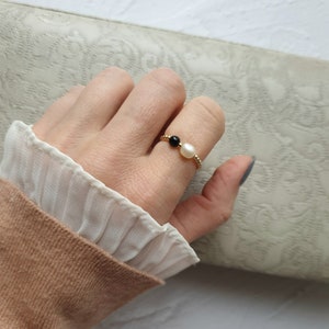 May include: A gold ring featuring a black bead and a white pearl. The ring is on a finger, with a tan sweater and white blouse in the background. The ring's band has a textured design.