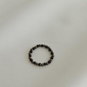 K&ouml;nnte beinhalten: Ein schwarzer und bronzefarbener Perlenring. Der Ring besteht aus kleinen, runden Perlen, die zu einem Kreis zusammengef&uuml;gt sind.
