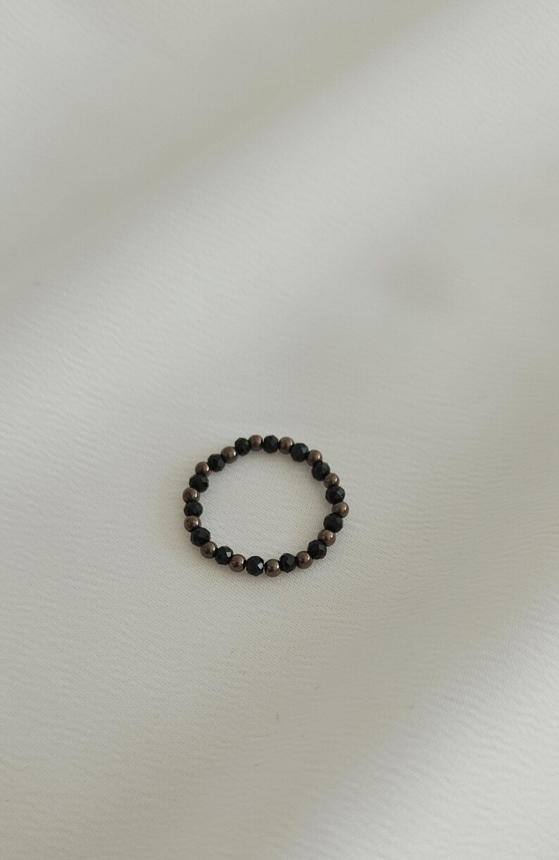 K&ouml;nnte beinhalten: Ein schwarzer und bronzefarbener Perlenring. Der Ring besteht aus kleinen, runden Perlen, die zu einem Kreis zusammengef&uuml;gt sind.