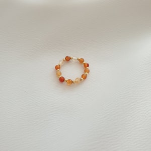 May include: A beaded ring featuring a stretch design. The ring is composed of small, round beads in shades of orange, gold, and clear. The beads are strung together with gold-colored spacers. The ring is displayed on a white background.