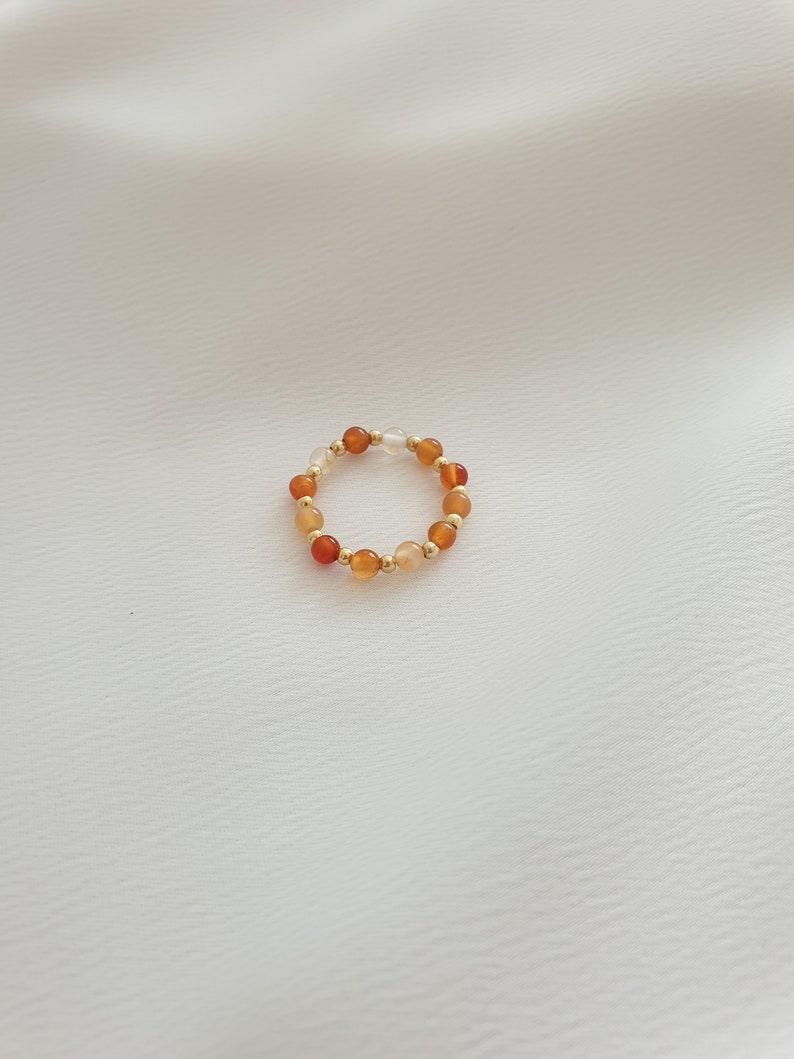 May include: A beaded ring featuring a stretch design. The ring is composed of small, round beads in shades of orange, gold, and clear. The beads are strung together with gold-colored spacers. The ring is displayed on a white background.