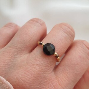May include: A gold beaded ring with a black gemstone. The ring is on a person's finger.
