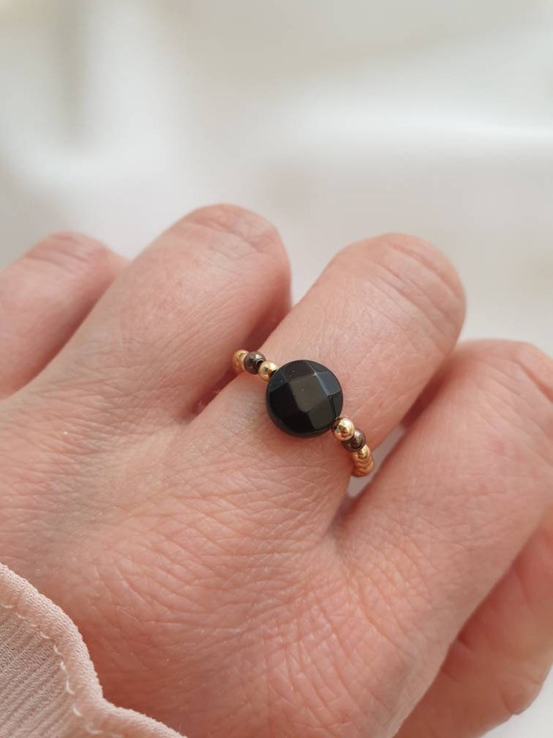 May include: A gold beaded ring with a black gemstone. The ring is on a person's finger.